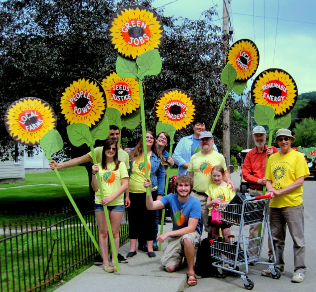 A smiling group of various individuals holding up colorful signs - local farms, people power, seeds of justice, community gardens, local power, renewable energy