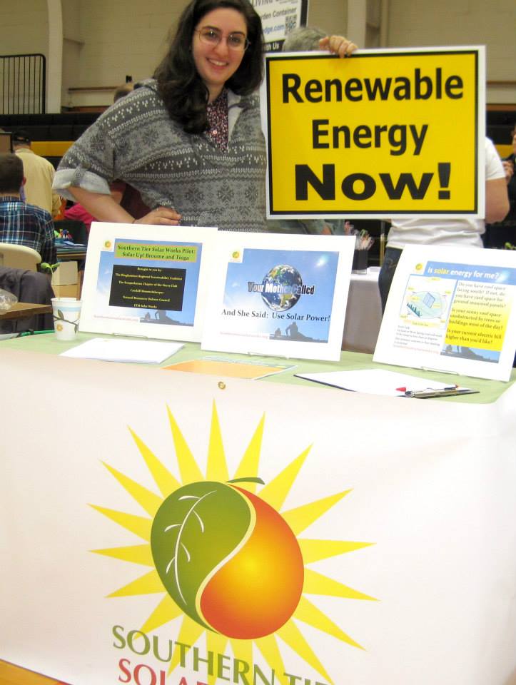 A table filled with information about solar power staffed by a friendly person holding up a sign that reads "renewable energy now!"