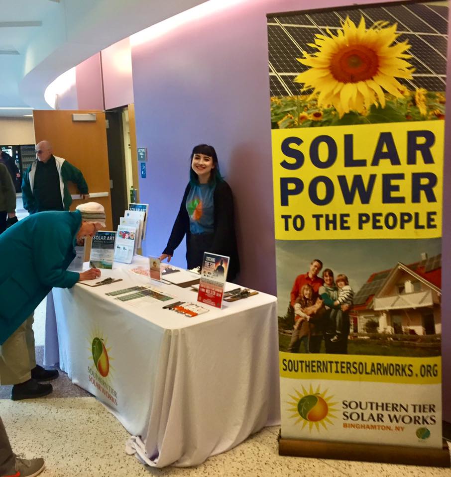 A smiling person standing behind a table filled with information about solar power and a person filling out a sheet on a clipboard